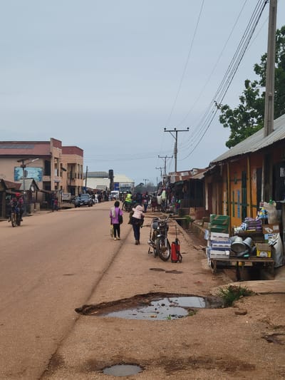 The image depicts a bustling street scene in Tangu, Nigeria, likely during the daytime under an overcast sky. The street is lined with various shops and buildings, suggesting a commercial area. On the right side of the street, there's a dusty patch of ground with puddles of water, possibly indicating recent rain.

Several people are visible, walking along the street and near the shops. Some individuals are carrying items, and a few are seen near motorcycles, which are common modes of transportation. One person in the foreground is wearing a pink top and black pants, while another is in a purple top. Further down the street, more people and a few vehicles, including trucks and cars, are present, indicating ongoing activity.

The shops appear to be selling a variety of goods, with items displayed outside. One shop features colorful painted doors and shelves stocked with what seem to be household items and provisions. The overall atmosphere suggests a lively marketplace or a busy residential street with commercial establishments.

There is some text visible on signage, including what appears to be "BARKA" on a blue poster featuring a man's face. Another sign with red lettering is partially obscured. The presence of power lines and poles overhead indicates infrastructure common in urban and semi-urban areas. The scene conveys a sense of everyday life and commerce in the community.