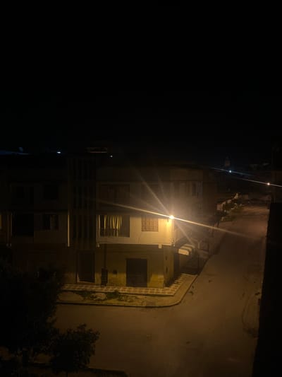 The image depicts a nighttime street corner in Ksar-el-Kebir, Morocco, viewed from an elevated vantage point. A multi-story building, likely residential or mixed-use, occupies the corner on the left. Its ground floor features two dark, closed doorways and light-colored, possibly yellow, rendered walls with a darker base. A patterned sidewalk precedes these entrances. The upper floor exhibits windows, one with light-colored curtains and another with closed brown shutters. Vertical slat-like architectural details are present on the upper left facade. The building's flat rooftop supports a satellite dish and a water heater or similar utility equipment.

A prominent streetlamp on the right side of the main building emits a strong, warm light that casts illumination onto the intersection and causes visible lens flares. The two intersecting streets appear dark and somewhat reflective under the artificial light. A dark tree or large bush partially occupies the lower left foreground. In the distant background, against the dark sky on the right horizon, a faintly illuminated minaret is visible, indicating the presence of a mosque. No individuals, vehicles, or active engagements are observable within the scene.
