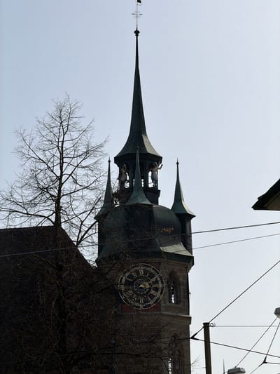 This image depicts an upward-angled view of the bell tower of St. Nicolas Cathedral in Fribourg - Freiburg, Switzerland, set against a bright, clear sky.

The prominent central subject is a tall, dark, multi-tiered clock tower. Its uppermost feature is a slender, pointed spire, crowned with a weather vane consisting of a small rectangular flag, a decorative cross-like element, and an arrow. Below the spire, a dark, multi-faceted roof structure encloses an open belfry section. Within this belfry, two anthropomorphic, automated figures are visible: one on the left in lighter attire, and one on the right in blue and white clothing with its right arm raised. Several smaller, dark spires are situated at various levels around the main tower structure.

Below the belfry, a large, ornate circular clock face is mounted on the stone tower. It features a dark background with prominent golden Roman numerals and intricate gilded detailing. The visible lower section of the tower is constructed from dark, rectangular stone blocks, and a recessed arched opening is visible to the right of the clock face.

To the left of the tower, a large, leafless deciduous tree with dark, intricate branches extends upwards, partially obscuring the lower-mid section of the tower and a dark building roofline in the lower-left corner. Multiple dark utility wires traverse the mid-ground and foreground horizontally and diagonally across the frame. A portion of a light-colored building's roof edge is visible in the upper-right corner. The overall lighting suggests bright daylight, resulting in some silhouetting of the darker tower and tree against the pale sky.