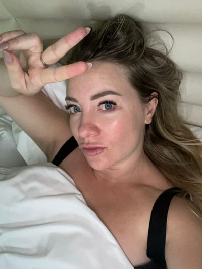 A woman lies on white bedding with her head tilted back. She makes a peace sign with her fingers. Her long, blonde hair is tousled. She has fair skin, blue eyes, and full lips. A black strap of fabric is visible near her shoulder. The background is out of focus, suggesting a close-up shot taken indoors. She is wearing a silver ring on her finger.