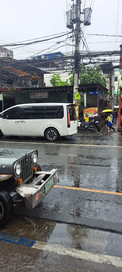 A rainy day in Mandaluyong City, Philippines is depicted in this vertical, eye-level shot. The scene unfolds along a street lined with buildings and an abundance of overhead electrical wires. In the foreground, the front of a vintage, olive-drab jeep is visible, its license plate reading "VAA 511". The jeep is parked on a wet asphalt road marked with a yellow line. Further down the street, a white van is parked. A group of pedestrians walks on the sidewalk, one person holding a yellow umbrella. A motorcycle is also parked near the sidewalk. Overhead, power lines are densely packed, supported by utility poles. A "CARWASH OPEN 24 HOURS" sign is prominently displayed on one of the buildings, and a street sign indicates "Agipag". The weather is overcast, and puddles on the road suggest recent or ongoing rainfall.