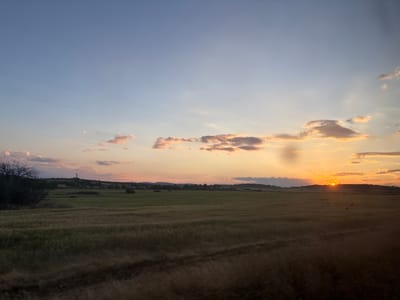 The image depicts a rural landscape at sunset, identified as near Smilets, Bulgaria. The sky occupies the upper two-thirds of the frame, transitioning from a clear light blue in the upper left to a gradient of pale yellow and orange near the horizon. Scattered cumulus clouds are present, with those closer to the horizon displaying orange and pink coloration from the setting sun. A faint, linear contrail is visible in the upper central sky. The setting sun is a bright, obscured orange orb positioned just above the distant tree line on the right side of the frame. Below the sky, expansive agricultural fields, primarily varying shades of green and brown, stretch towards a distant, undulating horizon of low hills or elevated terrain. Dark green foliage from trees and bushes is visible on the far left and sparsely along the distant horizon. Indistinct structures, possibly power poles or small buildings, are discernible on the distant hills. The immediate foreground shows a darker, slightly blurred section of the field, consistent with motion blur. No human figures or animals are present.