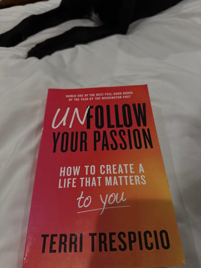An indoor scene in McAllen, United States of America, depicts a self-help book resting on white, rumpled bedding. The book, titled "UNFOLLOW YOUR PASSION: HOW TO CREATE A LIFE THAT MATTERS to you" by TERRI TRESPICIO, features a red to orange gradient cover. A banner at the top indicates it was "NAMED ONE OF THE BEST FEEL-GOOD BOOKS OF THE YEAR BY THE WASHINGTON POST". Behind the book, partially visible, are the hind legs and lower body of a black animal, likely a dog, resting on the same white bedding. The animal's fur appears dark with a possible small white marking. No people are visible. The environment consists solely of the soft, textured white fabric surface and the two main subjects.