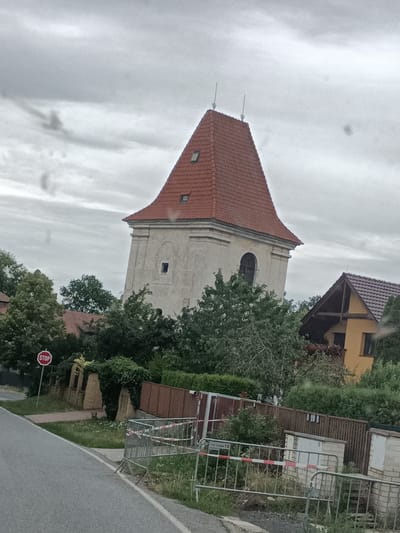 This media file captures an overcast day in Dřevčice, Czechia. The primary subject is a tall, pale building with a distinctive red, multi-pitched roof, resembling a tower or belfry, which appears to be leaning slightly. It has small, dark rectangular windows and a large arched window opening on one side. Adjacent to this structure, a smaller yellow building with a brown tiled roof is partially visible, suggesting a residential or utility area.
The scene is characterized by lush green trees and dense foliage surrounding the buildings. In the foreground, a grey asphalt road curves to the left, with a white line marking its edge. To the left of the road, a red circular STOP sign is mounted on a pole. A low stone wall, partially covered in ivy, runs along the edge of a property. Beyond the wall, a metal barricade with red and white striped tape is erected, possibly indicating a construction or restricted area. Some smaller brick structures and a wooden fence are also visible behind the barricade.
The sky is uniformly grey and cloudy, suggesting a dull or rainy weather condition. The lighting is diffused, without strong shadows, which is typical of an overcast day. There are some circular smudges on the surface of the image, likely due to water droplets on the camera lens.
Visible text includes the word "STOP" on the sign and numbers on some of the structures behind the barricade, including "50" and what appears to be "201YDAM 50" or similar text. The overall impression is one of a quiet, possibly rural or suburban, setting on a gloomy day. No people are visible in the frame.