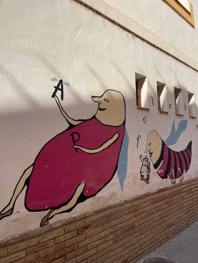 A vibrant mural adorns a textured, off-white building wall in Picanya, Spain, depicting two large, cartoon-like figures under clear, sunny conditions. Strong shadows are cast from the left, highlighting the wall's uneven surface and architectural features.

The larger, bottom figure is upright, rendered in a light skin tone with black outlines, and wears a flowing magenta dress or tunic. It has an elongated neck and appears to be looking down at its left hand, which holds a stylized black letter "A" with two light blue, wing-like accents. On its chest, a small, black 'P' or 'p' is visible.

Above and slightly to the right, a second, smaller figure is painted upside down. This figure also features light skin and black outlines, wearing a striped, conical cap and a matching striped outfit in shades of dark red/maroon and black. Its face shows a subtle, content expression. Small, abstract black symbols resembling letters like 'f' and 'JL' are scattered around this figure's hands and head.

To the left of the upper figure, a series of five rectangular openings or small windows are cut into the wall, casting their own distinct shadows. Adjacent to these openings are faint, hand-drawn black characters, including what appears to be 'u', 'N', and other indistinct marks, possibly indicating a sundial or a sequence. A narrow, vertical groove or line runs alongside these openings, further suggesting a gnomon.

On the far right, a section of a brick wall, composed of reddish-brown bricks, runs perpendicular to the painted wall, suggesting an corner or an adjacent structure. The overall scene is whimsical, with the mural adding a playful and artistic touch to the building's exterior.