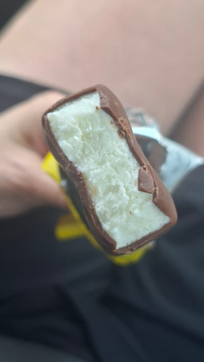 A close-up, eye-level shot shows a chocolate-covered ice cream bar held by a person's hand in Лугаваслабадскі сельскі Савет, Belarus. The ice cream is white and appears to have a slightly frosty texture, indicating it is cold. The chocolate coating is dark brown and has a few small chips and imperfections, suggesting it has been bitten into. A portion of bright yellow wrapper is visible beneath the ice cream bar, peeking out from the bottom left of the frame. The background is out of focus, with hints of blurred skin tones and dark fabric, suggesting the setting is likely outdoors or in a casual indoor environment. The lighting is moderate, casting soft shadows and highlighting the textures of the ice cream and chocolate.