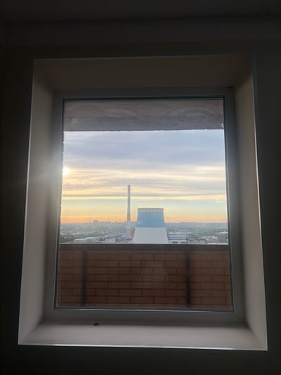 This image depicts a view through a window overlooking a city, likely Shlisselburg, Russia, with a prominent cooling tower in the middle ground. The scene is captured during what appears to be sunrise or sunset, with the sun low on the horizon, casting a warm glow across the sky and illuminating some of the buildings.

The foreground is dominated by the window frame, which is painted white. Below the window pane, there is a brick wall, suggesting the view is from an elevated position. The brickwork is laid in a stretcher bond pattern.

In the middle ground, a large, white and light blue cooling tower stands out. It has a wide base that tapers towards the top, which is rimmed with a decorative pattern. To the left of the cooling tower, a tall, slender smokestack pierces the sky. The rest of the city stretches out behind these structures, a dense collection of buildings of varying heights, some appearing to be industrial and others residential. The sky above is filled with soft clouds, colored in hues of orange, pink, and pale blue, indicative of the time of day. The overall impression is one of an industrial urban landscape under a tranquil sky.