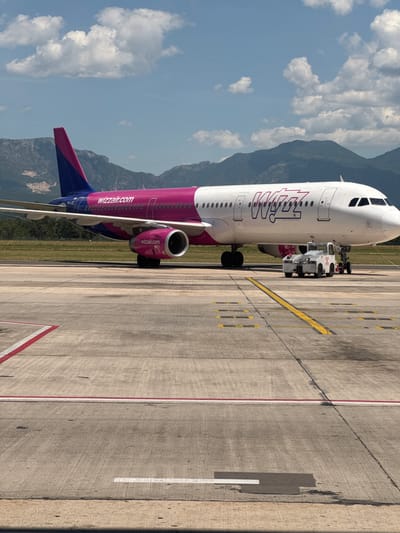 A Wizz Air airplane with the registration HA-LIC is parked on an airport tarmac. The aircraft is predominantly white with a pink stripe along the fuselage and pink and blue accents on the tail. The Wizz Air logo is prominently displayed on the side of the plane. In front of the aircraft, a white tug vehicle is positioned, suggesting the plane is either being prepared for departure or has recently arrived. The background consists of a mountainous landscape under a partly cloudy blue sky. The foreground shows the concrete surface of the tarmac, marked with yellow and red lines indicating taxiways and runways. The scene appears to be at Tirana International Airport (TIA), which serves the capital city of Tirana and is located approximately 25 kilometers (16 miles) northwest of Kruja, Albania.