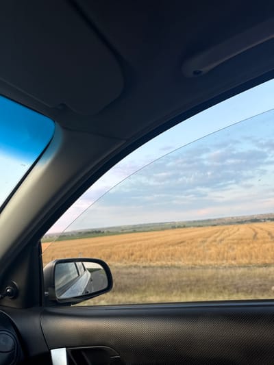 The image provides a view from inside a vehicle, looking out through the side window. The perspective suggests a journey through a rural landscape.

In the foreground, the interior of the car is visible. The dark textured material of the car door panel dominates the lower right portion of the frame. A black car door handle is also visible. To the left, a rearview mirror is mounted on the car's A-pillar. The mirror reflects the road and the side of the car, showing the text "OBJECTS IN MIRROR ARE CLOSER THAN THEY APPEAR". Part of the dashboard and the car's headliner are visible along the top edge of the image.

The main subject of the exterior view is a vast field of golden, harvested crops, likely wheat or barley, stretching towards the horizon. Tractor tire tracks are clearly visible in the field, indicating recent agricultural activity. Beyond the field, a gentle slope of land is visible, with scattered vegetation and possibly some distant structures. The sky above is a pale blue, dotted with scattered, wispy clouds, suggesting it is either early morning or late afternoon.

The scene is consistent with the rural outskirts of Sadaclia, Moldova, a region known for its agricultural landscape, particularly grain cultivation. The image captures a moment of transit, emphasizing the open, cultivated terrain typical of this area. There are no people or animals present in the image. The lighting and atmospheric conditions suggest a mild, possibly overcast, day.