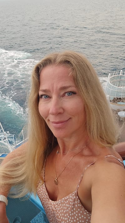 A woman with blonde hair and blue eyes is pictured in a selfie taken on a boat on the sea. She is wearing a polka-dot dress and a gold necklace. The location is assumed to be on the sea, as the boat is moving and the water can be seen. The city location of Unknown, France cannot be confirmed due to the photograph not showing enough identifiable clues.