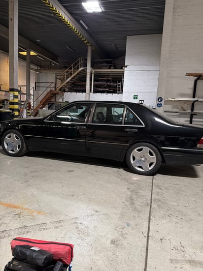 A black Mercedes-Benz S-Class sedan is parked in a dimly lit industrial garage. The car is in profile, facing right. The garage has a concrete floor with visible seams and some scuff marks. Above, the ceiling is constructed of corrugated metal and industrial beams, with yellow and black hazard stripes visible on one beam. Fluorescent lights provide illumination. To the left, a concrete pillar with yellow and black stripes supports the ceiling. In the background, there is a metal staircase leading up to a platform, and white brick walls are visible. A green "Ausgang" (Exit) sign is mounted on the wall. On the floor in the foreground, a red and black tool bag lies next to other dark objects. The overall impression is a utilitarian, possibly workshop or storage space. The image does not contain enough information to determine the specific location within Osnabrück, Germany.