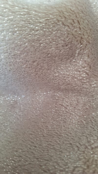 The image shows a close-up, abstract view of a soft, plush fabric with embedded glittery specks. The fabric is a muted, light brown or beige color. The texture appears fluffy and possibly synthetic, with visible fibers and a slightly uneven surface. The glitter specks are small and scattered, catching the light and creating a subtle shimmer. The image is out of focus, emphasizing the texture and light play rather than any specific details. The scene has no identifiable context related to the city of Nogliki, Russia, or any specific objects, people, or actions.