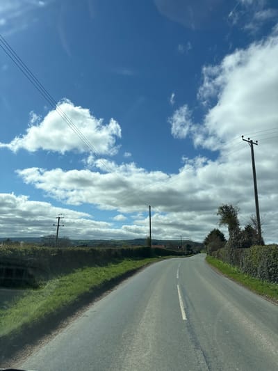 The image shows a rural road in Longden, United Kingdom. The road is asphalt, with a broken white line marking the center. Verges of grass and hedgerows line both sides of the road. Overhead power lines are visible, supported by wooden poles. The sky is blue with scattered white clouds. No people are visible.