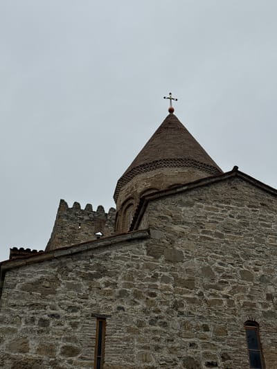 Exterior shot of the Svetitskhoveli Cathedral, located in the historic city of Mtskheta, Georgia. The architecture showcases stone masonry in light gray tones. The frame emphasizes the church's pointed tower capped with a cross, viewed against an overcast sky. Details include windows, roof edges and decorative masonry patterns, all suggesting the cathedral's historical significance and construction details.