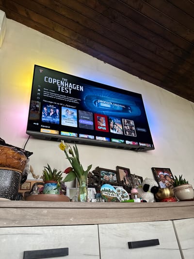 The image is of a television mounted on a wall in what appears to be an indoor setting. The TV is displaying a show titled "The Copenhagen Test," with various menu options visible on the screen. Adjacent to the TV, on the right, there's a collection of decorative items, including framed pictures, potted plants, and figurines placed on a shelf or ledge. To the left of the TV, the wall is paneled with dark, wooden planks. The scene appears to be indoors. The exact location or interaction cannot be discerned from the image. The OCR data suggests that the language on the TV screen is Bulgarian, which indicates Lipnik, Bulgaria, but there is no information about the city of Lipnik to confirm it is the location of the image.