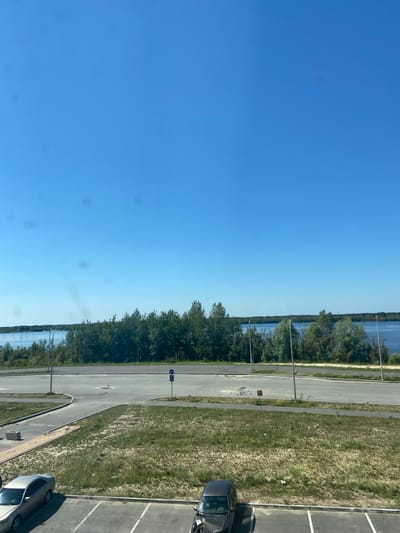 An aerial view captures a clear, cloudless blue sky above a serene landscape. In the middle ground, a dense line of green trees separates a paved area from a tranquil body of water, likely the Ob River, as the image is geotagged to Nefteyugansk, Russia. The paved area appears to be a road or parking lot, with a traffic sign visible. In the foreground, a parking lot with white markings is partially visible, with two cars parked: a silver sedan on the left and a dark-colored hatchback on the right. The ground is a mixture of asphalt and dry, grassy patches.