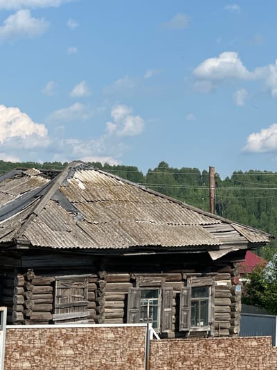 The image shows an old, weathered log house with a damaged, corrugated roof under a blue sky with scattered clouds. Several rows of logs form the walls, and there are two windows visible with wooden shutters, one of which is partially boarded up. A fence with a brick-like pattern is in the foreground. In the background, a dense green forest stretches across a hillside, with a wooden utility pole and power lines in front of it. A small blue sign with the number "12" is attached to the corner of the house. The scene is likely a residential area in the village of Yelzovka, Russia, given the traditional log construction and rural setting. There are no people visible in the image.