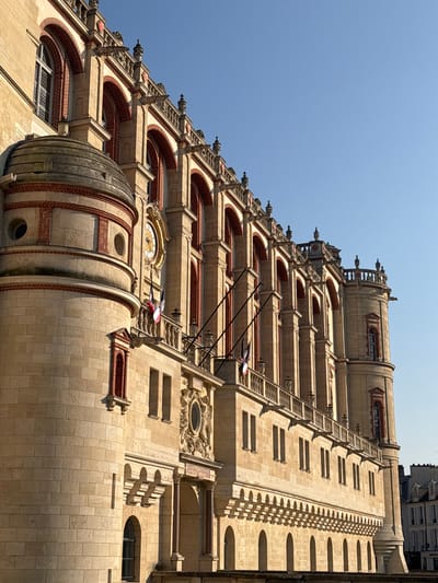 This image presents a forensic view of the Château de Saint-Germain-en-Laye, located in Saint-Germain-en-Laye, France. The scene is illuminated by direct sunlight under a clear, cloudless blue sky, creating strong contrasts and distinct shadows on the building's facade.
The structure is predominantly composed of light-colored cut stone blocks, with architectural detailing highlighted by red brickwork around arches, windows, and decorative banding. On the far left, a substantial circular tower features a domed roof and several small, round window openings, alongside a rectangular window framed in red brick.
The main facade extends horizontally, showcasing multiple architectural levels. The upper section features a series of evenly spaced arched windows, each framed in red brick, surmounted by a decorative stone balustrade adorned with ornate finials and grotesque figures. Below this, a golden clock face with black Roman numerals is centrally positioned within the stone masonry. Beneath the clock and extending along a significant portion of the facade is a stone balustrade, from which three French national flags (blue, white, red vertical stripes) are displayed diagonally on poles. Further down, multiple rows of rectangular windows are visible, and the lowest exposed level of the building exhibits a series of smaller, regularly spaced arched windows.
On the far right, a smaller, circular turret-like structure, consistent with the main building's materials and style, features narrow, arched windows. No individuals or specific actions are depicted within the frame. In the distant lower right background, a portion of another urban building with multiple windows and a dark roofline is visible, indicating an established urban setting adjacent to the château.