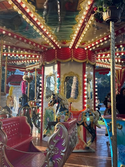 The image displays an illuminated, ornate carousel located outdoors in the Prato di Arezzo, Arezzo, Italy. The scene is bathed in warm, directional sunlight, indicating late afternoon or early evening. The carousel features a multi-tiered structure with a decorated ceiling reflecting numerous small, white lights that outline red and gold trim and draped red fabric. Ornate golden-framed mirrors are embedded into the central panels, reflecting surrounding trees and light. The carousel figures include multiple classic horses, such as a black horse with gold, blue, and orange accents prominently featured on the right, and lighter-colored horses with pink and yellow plumes visible on the left. An ornate red and gold chariot or bench occupies the immediate foreground. Three individuals are visible: one person in a light jacket is seated on a lighter-colored carousel horse on the left; another person in dark attire is partially obscured behind a dark carousel horse; and a woman with dark hair and dark clothing is positioned on the far right, near another carousel figure. The carousel is set on a paved or wooden surface, with mature trees visible in the background, characteristic of a park environment.