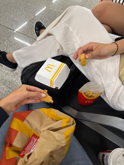 The image depicts an indoor scene at Palma de Mallorca Airport in Son Santjoan, Spain. Two individuals are present, consuming fast food. The person on the left wears blue jeans and holds a partially eaten food item, consistent with a burger or sandwich, in their right hand. A brown McDonald's paper bag, containing a red ketchup packet, rests adjacent to this individual. The person on the right, dressed in white wide-leg trousers and black sneakers, holds a golden-brown fried item, resembling a chicken nugget or onion ring, in their right hand. A black suitcase is positioned between the two, supporting a white McDonald's container box and a red McDonald's french fry carton filled with fries. A white cup is partially visible on the floor to the right. The flooring consists of light grey tiles with distinct white grout lines. A third individual's upper thigh is visible in the upper right background.