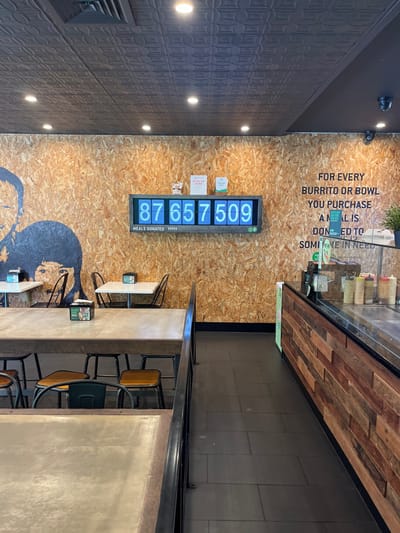 The image displays the interior of a fast-casual restaurant or takeaway, possibly in Kelvin Grove, Australia, seen from a slightly elevated angle. The space appears empty of customers or staff.
The dominant feature is a light brown, raw wood-textured (possibly OSB) wall at the back, adorned with a large digital display. This display prominently shows "87,657,509" in blue digits, with smaller text beneath reading "MEALS DONATED". A faint, smaller number "39999" is also visible below the "Meals Donated" text. Adjacent to the digital counter, a small white sign indicates "PICK UP HERE," and another smaller green sign is partially visible.
Above the counter, a clear message is stenciled in large black letters on the wooden wall: "FOR EVERY BURRITO OR BOWL YOU PURCHASE A MEAL IS DONATED TO SOMEONE IN NEED." Below the digital display, a large, stylized black graphic depicting a person's profile is painted on the wall.
To the right, a long service counter features a facade of dark wooden planks and a glass display case on top, likely for ingredients or condiments. The floor in this service area is covered with dark, rectangular tiles.
The dining area in the foreground and middle ground consists of several light-colored rectangular tables paired with industrial-style chairs. These chairs have black metal frames and seats in varying shades of brown and dark green.
The left wall of the establishment is dark, featuring a patterned or textured surface, and is illuminated by multiple recessed circular light fixtures. The overall aesthetic is modern and functional, highlighting the establishment's charitable initiative.