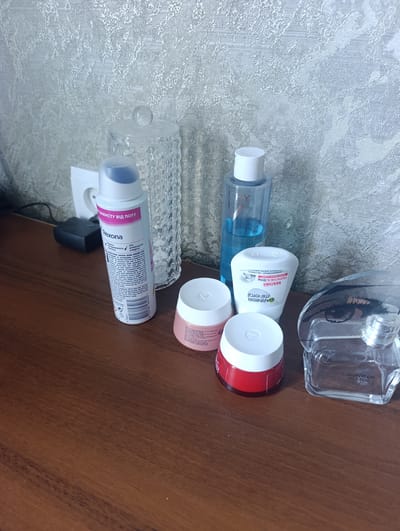 In Nikopol, Ukraine, on a wooden surface, a collection of cosmetic products is displayed. From left to right, there is a white aerosol deodorant can with pink lettering that reads "Rexona," a clear glass jar with a patterned design, a blue-capped bottle of Vichy toner, a small pink jar with a white lid, a red jar with a white lid, a white stick deodorant for Garnier, and a clear glass perfume bottle with "CALVIN KLEIN WOMEN" visible on it. A white electrical outlet and a black adapter are also visible on the wall behind the items. The background is a textured, light-colored wallpaper.
