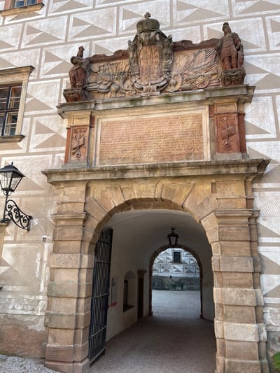 The image shows an ornate stone archway leading into a courtyard. Above the arch, a large stone inscription plaque is mounted on the building's facade, framed by carved decorative elements. The building's exterior is adorned with a geometric pattern of beige and light brown triangles. To the left of the arch, a black wrought-iron lamppost with a glass lantern is attached to the wall.
The archway itself is constructed of large, rough-hewn stone blocks. A dark metal grate is partially open on the left side of the arch, allowing a view into a dimly lit passageway. Inside the passageway, a hanging lantern is visible. The courtyard beyond the archway features more buildings with similar geometric facade patterns and a gravel ground.
This scene is the entrance to the Náchod Castle in Náchod, Czechia. The inscription plaque above the archway is a historical marker, likely commemorating a significant event or person related to the castle's history. The architectural style suggests a Renaissance or Baroque influence, common in Central European castles.