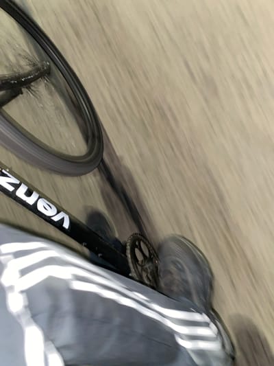 A blurry image depicts a low-angle view of a moving bicycle on a paved surface, possibly in Monte Grande, Argentina. The bicycle frame, partially visible, is black with white lettering "VENZ". A wheel spins on the left side, and the chainring is also visible. Adjacent to the bicycle is a person wearing gray pants with white stripes on the side. The surrounding environment appears to be a street or walkway with indistinct surface details due to the motion blur.