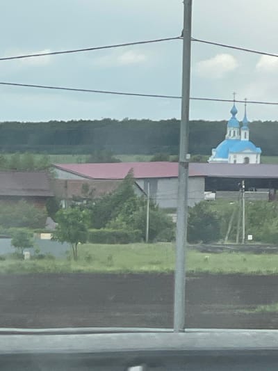 The image, shot through a car window frame, shows a distant view of what appears to be an Orthodox church with blue domes. The architecture is partially obscured by trees and other buildings. Power lines are visible in the foreground. The landscape surrounding the church appears to be rural, with trees and open fields. The location is Кокино, Russia.
