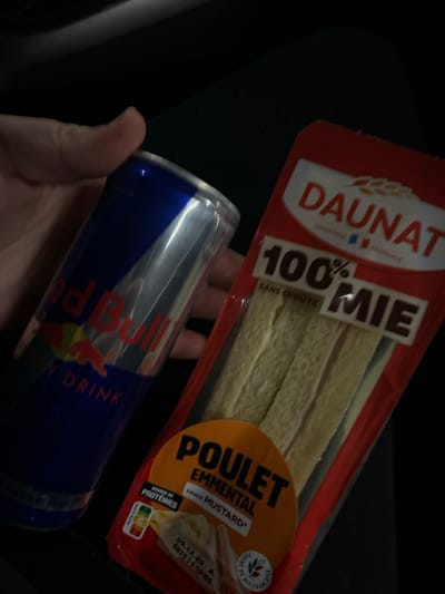 An overhead, close-up image depicts a light-skinned hand holding a Red Bull Energy Drink can and a packaged food item against a dark, indistinct background. The Red Bull can is blue and silver, displaying its prominent logo. To the right, the packaged food item, branded "DAUNAT," is visible in a red plastic wrapper. It contains three pale, rectangular bread-like segments labeled "100% MIE SANS CROUTE" (100% soft bread without crust). An orange circular label on the package indicates the filling: "POULET EMMENTAL sauce MUSTARD" (Chicken Emmental mustard sauce), alongside a "SOURCE DE PROTÉINES" (Source of Protein) label and a Nutri-Score F rating. A partial date, "09.12.23," is visible, likely indicating an expiration date. The scene is located in the city of Andermatt, Switzerland.