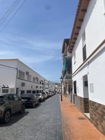 The image depicts a narrow street in Pizarra, Spain, lined with whitewashed buildings. The street is paved with dark grey cobblestones on the left side, where several cars are parked, and a reddish-brown paved walkway on the right side. The buildings on the left are two stories high, with numerous windows and air conditioning units visible. The building on the right is also two stories high, with a tiled roof and balconies. The sky is clear blue with some wispy clouds. In the distance, the street curves and leads towards more buildings and a church steeple. There are no people visible in the image. The scene suggests a quiet, residential or commercial street in a Spanish town on a sunny day.