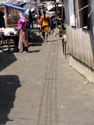 The image captures an outdoor scene on an unpaved street or alleyway in Oworonshoki, Nigeria, characterized by informal market activity and residential structures. The ground is dirt-packed, showing scattered debris and refuse. Strong sunlight from an overhead source casts distinct, elongated shadows, including those of individuals and thin linear patterns, possibly from overhead wiring or supports.
On the left, a woman stands with her back largely to the viewer, wearing a vibrant pink short-sleeved top, a multi-patterned brown, orange, and blue wrap skirt, and a white and blue patterned headscarf. She is positioned beside a makeshift wooden stall or table displaying various items, some in plastic containers or bowls, beneath a blue tarp awning.
Further into the scene, centrally located, another woman walks away from the camera, dressed in a yellow and orange patterned short dress. To her immediate left, a young boy in a red shirt walks in the same direction. In the midground and background, multiple individuals are discernible, moving or standing amongst a dense cluster of rudimentary structures, many appearing to be shacks or stalls constructed from corrugated metal sheets and covered with tarpaulins.
The right side of the frame is dominated by the rough, vertically paneled wall of a structure, likely corrugated metal, with a concrete base. A small, open-shuttered window is visible on this wall, and a simple wooden table or stand is positioned in front of it. The overall setting indicates a lively, unformalized urban or semi-urban environment.