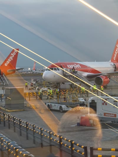 This image depicts an airport scene at dusk in Somma Lombardo, Italy, likely at Milan Malpensa Airport (MXP). Two EasyJet airplanes are visible on the tarmac. The airplane in the foreground has its front passenger door open, with a set of stairs leading up to it. Several individuals wearing high-visibility vests are seen near the airplane, some appearing to be ground crew. A baggage tug and other airport service vehicles are also present. The background shows the lights of another EasyJet airplane and the distant city skyline. The lighting suggests a transition from daylight to night, with artificial lights from the airport beginning to illuminate the scene.