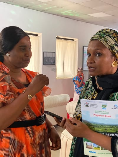 Two women are in conversation, possibly at an event. The woman on the right, wearing a patterned headscarf and gold earrings, holds a program titled "Agro-Climatic Resilience in Semi-Arid Landscapes: Program of Event" with logos for the Federal Government of Nigeria and the World Bank. The other woman, in an orange dress with ruffle details and a black belt, looks up at her. She is wearing earrings and bracelets. A phone is visible in the woman's hand on the right. Two framed pictures are affixed to the wall behind the women. Based on the program documentation, the setting is possibly in the city of Kwarau, Nigeria, given the Federal Government of Nigeria and landscape imagery.