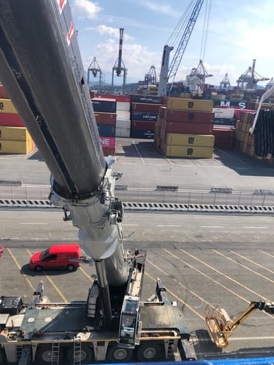 The image displays a container terminal in La Spezia, Italy, under bright daytime conditions. In the foreground, a large, dark gray Liebherr mobile crane, with its telescopic boom partially extended and angled upwards, dominates the lower left-to-mid frame. A single person is discernible inside the crane's operator cab. To the left of the crane, a red commercial van is parked on the asphalt ground, which features yellow painted lines and numerical markings. On the far right, the yellow platform cage of an aerial work platform is partially visible. The midground reveals an expansive container yard filled with numerous shipping containers stacked several high. These containers exhibit various colors including red, yellow, blue, and white, with visible corporate branding from companies such as MSC, CMA CGM, and ONE. The background features multiple large gantry cranes typical of a port environment, with distant hills or mountains visible under a blue sky with scattered white clouds. The overall scene is static, depicting industrial port operations.