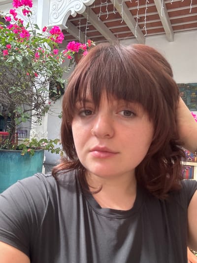 A woman with brown hair and bangs poses for a selfie. She wears a dark gray t-shirt and has a nose ring. Her face is slightly flushed, and she looks directly at the camera. In the background, a vibrant pink bougainvillea bush stands out against a backdrop of a building with white trim and a terracotta tiled roof adorned with string lights.