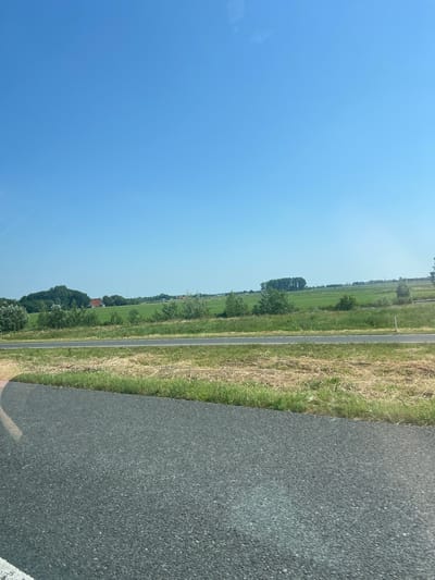 This is a landscape image taken in Marsum, Netherlands, on a clear, sunny day. The perspective is from the side of a road, looking out towards a rural scene. The image is oriented vertically, with the sky occupying the left side and the road the right.

The sky is a vibrant, cloudless blue, suggesting it is daytime, likely mid-morning or afternoon given the brightness. To the right of the sky, a narrow paved road runs vertically through the frame. The road appears to be asphalt, with a white line marking the edge.

Adjacent to the road, on the left side of the frame, is a grassy field. This field is a mix of green grass and drier, straw-colored vegetation, indicating a warm season. Further into the field, there are trees and some buildings, including what appears to be a red-roofed house. In the distance, a few windmills are visible, a common sight in the Dutch landscape. The overall scene depicts a tranquil rural environment.

There are no people or overt activities visible in the image. The focus is on the natural landscape and the road. The image conveys a sense of peace and openness. There is no visible text in the image.