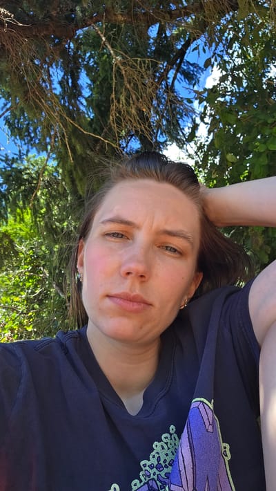A woman is taking a selfie outdoors in what appears to be a park or wooded area. She has her hand behind her head and is looking at the camera with a slightly pensive expression. The background is filled with lush green foliage and branches of trees, with patches of blue sky visible through the leaves. She is wearing a dark blue t-shirt with a graphic design on the front. The lighting suggests it is daytime, likely a sunny day, with dappled light filtering through the trees. The exact location in Cherry Grove, United States, cannot be determined from the image.