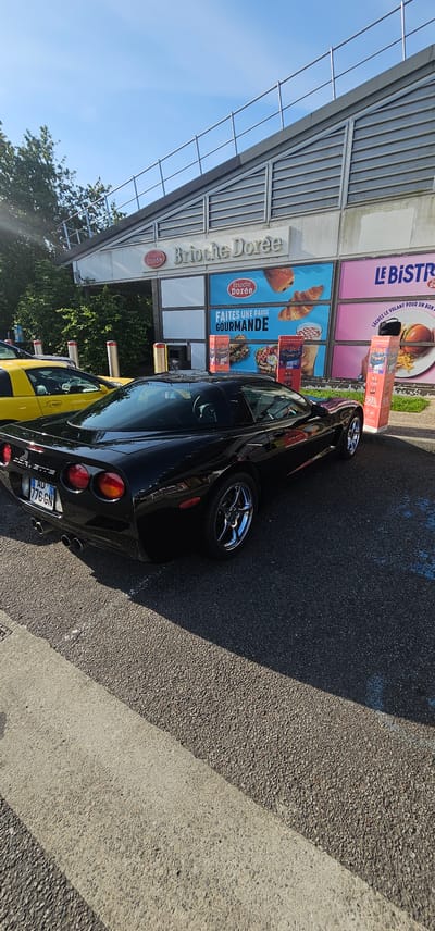 The image depicts an outdoor scene at the Aire de la Crèperie, a highway service area located in Bain-de-Bretagne, France.

In the foreground, a black Chevrolet Corvette C5, identifiable by its distinctive rear taillights and body shape, is parked on asphalt pavement. The car displays a French license plate reading "AD-776-GN" and features chrome multi-spoke wheels and dual exhaust pipes. A significant shadow from the adjacent building extends across the front half of the car and the ground. Partially visible behind the black Corvette is a yellow sports car, also appearing to be a Chevrolet Corvette, parked further forward.

The commercial building in the background features a sloped, grey, louvered roofline and large glass window panels. Prominent signage on the building reads "Brioche Dorée" and "LE BISTROT" in large lettering. Advertising panels on the windows display images of various food items, including croissants, pastries, burgers, fries, and salads, accompanied by French text such as "FAITES UNE PAUSE GOURMANDE" (Take a gourmet break) and "LÂCHEZ LE VOLANT POUR UN BON PLAT !" (Drop the wheel for a good dish!). Red and white striped bollards are positioned along the front of the building. A small black bag rests on top of one of the red promotional pedestals near the black car.

The environment includes green trees and foliage visible behind the yellow car and to the left of the building. The sky above is clear blue with minimal cloud cover, indicating a bright, sunny day. The pavement is asphalt, with a lighter concrete strip visible in the lower left corner. No people are visible in the scene.