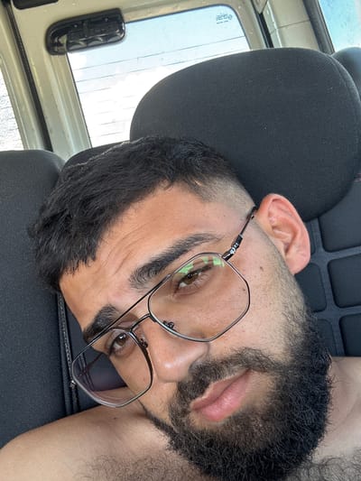 A man is taking a selfie in a vehicle. The man has dark hair, a beard, and is wearing glasses. The background shows a car interior with a headrest and a window. The lighting suggests it is daytime. It is not possible to determine the location in the city of Petrovo, Bulgaria from the image.