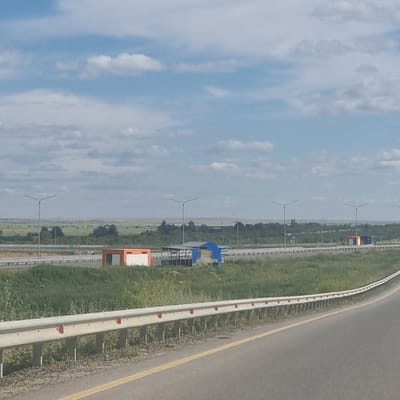 The image shows a highway scene under a cloudy blue sky, likely taken from a moving vehicle. In the foreground, the edge of a paved road is visible with a yellow lane marker. A metallic guardrail with red reflectors runs along the side of the road. Beyond the guardrail, a strip of green grass and vegetation separates the road from the main area.
In the middle ground, there are several structures and infrastructure elements. A long, white guardrail is present on the opposite side of the highway. Two small, orange and white modular buildings are situated near the highway. One appears to be a simple rectangular structure with a white door or panel. Another structure, partially covered with blue tarpaulin and green material, is next to it, resembling an open-sided shelter or construction area with some visible posts and beams. Several streetlights with a modern design are spaced along the highway.
The background depicts a vast, flat landscape with green fields and scattered trees, suggesting a rural or semi-rural environment. The horizon is low, and the sky is filled with scattered white clouds against a blue backdrop. The overall impression is one of an open, developed highway in a predominantly natural, undeveloped setting. The location is identified as Alğa, Kazakhstan.