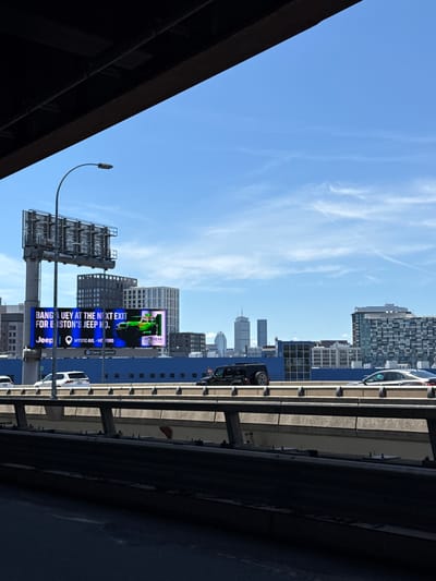 The image depicts an urban scene in Somerville, Massachusetts, USA. A highway overpass dominates the right side, with cars visible driving on it. A view of the city is visible, including buildings and skyscrapers. A large billboard is prominently displayed on the left side of the image, advertising Jeep and Ford. The sky is blue with wispy clouds. A street light and utility poles are also present in the view.