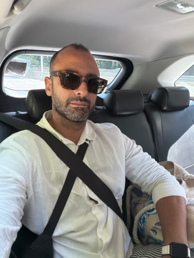 A man with a beard and sunglasses is sitting in the back seat of a car, wearing a white linen shirt and a seatbelt. He is looking at the camera. Outside the car, trees and a road are visible. The interior of the car is dark, with black leather seats. There is a bag with a nautical print on the seat next to him. The image was taken on July 22, 2023, in Cardano al Campo, Italy.