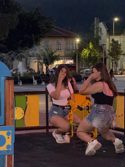 The image shows two young women on a playground at dusk in Debelets, Bulgaria. The woman in the foreground is wearing a pink t-shirt and denim shorts. She is sitting on a yellow and green playground structure and has her hands covering her face. The second woman, in the background, is wearing a black tank top and denim shorts. She is also on the playground equipment, leaning back with her hands covering her face. Both women appear to be in their late teens or early twenties. The playground features colorful structures, and behind it, there are buildings with illuminated windows and trees. A fountain is visible in the background, its water spraying upwards. The lighting suggests it is evening, with artificial lights from street lamps and buildings illuminating the scene. The overall mood seems playful and candid, possibly capturing a moment of silliness or lightheartedness between the friends. No visible text is present in the image, except for the word "СТОКИ" (meaning "goods" in Bulgarian) on a sign in the background, possibly indicating a nearby shop.