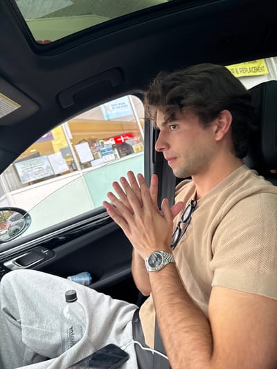A young adult male is depicted from the torso up, seated in the passenger side of a vehicle, gazing out the window towards a gas station convenience store. He has dark, wavy hair and is wearing a beige short-sleeved polo shirt, with black-framed eyeglasses resting on his collar. His left wrist displays a silver-colored watch with a Jubilee bracelet and a dark dial. His hands are clasped together in front of him, fingers interlocking. His legs, partially visible, are clad in light grey sweatpants, with an Essentia Water bottle and a black smartphone (connected by a white cable) resting on them. The vehicle's interior features dark upholstery, a visible sunroof, and a partially open window revealing the side mirror.
The external environment, viewed through the vehicle window, shows a Mobil branded gas station convenience store. Prominent signs visible include a blue banner stating "SAVE MORE AT THE PUMP," and a yellow sign above it with text related to "AUTOMOTIVE" and "REPAIR & REPLACEMENT." The Mobil logo (red horse on a blue background) is discernible on another sign. The convenience store interior displays shelving with various products.
The scene is located at the Mobil gas station at **787 Main Street, Waltham, Massachusetts, United States.**
