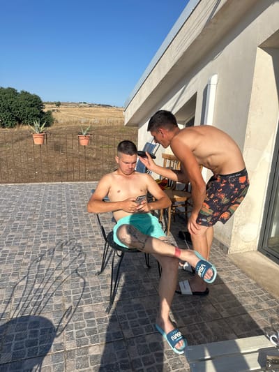 Two young men are on a paved patio area outdoors. The man on the left is bending over, his head tilted down as if attending to the other man. He is wearing patterned swim shorts. The man on the right is seated, leaning back and looking at his phone. He is wearing light blue swim shorts. Both men are shirtless and appear to be young adults.
The setting is bright and sunny, suggesting it is daytime. The ground is paved with gray cobblestones. Behind the men, there is a building with light-colored walls and a glass door or window. To the left of the building, there is a clear blue sky. To the right of the patio, there is a dry, grassy field with some scattered bushes and trees. Two potted plants are visible near the edge of the patio. The overall atmosphere seems relaxed and casual.
There are some visible brand names. The swim shorts of the man on the left have "Calvin Klein" written on the waistband. The slides worn by the man on the right have "VSVN" written on them. The image appears to be taken in Ragusa, Italy.