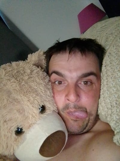 The image is a close-up, selfie-style photograph featuring a person lying down, partially obscured by a large teddy bear and a textured blanket or pillow. The person's face is angled towards the camera, with their tongue sticking out. They have brown eyes, dark hair, and stubble. The teddy bear is a light brown, plush toy with visible eyes and a snout. The background includes a hint of a pink lampshade, suggesting an indoor setting, possibly a bedroom. The lighting appears to be dim, indicating it might be nighttime or early morning. The overall mood conveyed is playful or silly, due to the tongue-out pose and the presence of the teddy bear. No visible text is present in the image. The location context provided is Wedel, Germany.