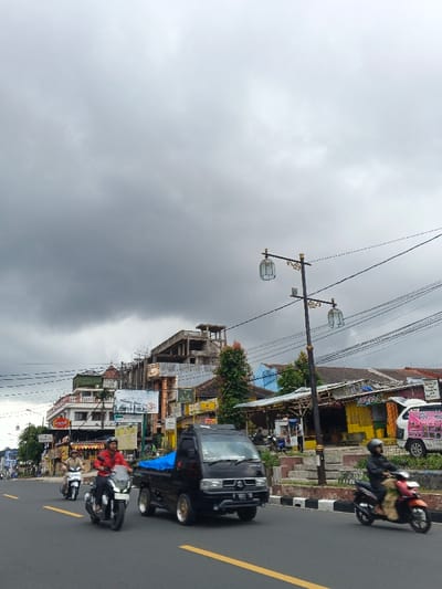 An urban street scene in Palayan, Philippines, is depicted under an overcast sky. A multi-lane asphalt road with yellow center lines and white solid lines occupies the foreground. Multiple vehicles are in motion: a black utility pickup truck with a blue tarp in its bed is central, moving right. Ahead of it, a person wearing a red shirt and black pants drives a dark gray scooter. Behind the truck and further left, another person rides a silver scooter. To the right of the truck, a third person, wearing dark clothing and a black helmet, rides a red-orange scooter.
The background features a densely packed array of buildings. On the left, multi-story structures with balconies and commercial signage, including one sign for "HOTEL PARADISE," line the street. Prominently in the mid-background is a multi-story concrete structure with exposed rebar, indicative of ongoing construction or an unfinished building. To the right, lower-rise, more informal structures, some painted yellow and blue, appear to be shops or stalls. A black streetlamp post with two decorative white lanterns stands on a raised median or sidewalk on the right side of the road. Numerous overhead electrical wires crisscross the upper mid-ground. Green foliage is visible between some buildings and near the streetlamp. Concrete barriers separate the road from the sidewalk/median.