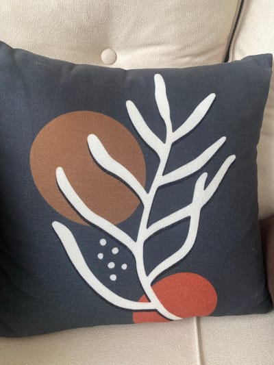 A dark blue pillow with a white abstract branch design and scattered brown and rust-colored circles is placed on a light-colored textured surface. The pillow is square and appears to be made of fabric. The design on the pillow is modern and minimalist. The background surface is likely a couch or a similar piece of furniture, indicated by the texture and the presence of a button detail on the left side of the image. The lighting suggests it is daytime. There is no visible text in the image. The setting appears to be indoors.