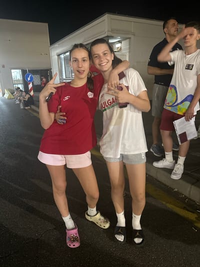Two young women are posing for a photo outdoors at night. The woman on the left is wearing a red t-shirt and light pink shorts, and she has her tongue sticking out and is making a peace sign with her fingers. The woman on the right is wearing a white t-shirt with "BULGARIA BASKETBALL" printed on it and grey shorts. She has her arm around the woman on the left and is holding a phone. Both women are wearing white socks and sandals. In the background, there are other people and a building that appears to be a border control station. The scene is dimly lit by artificial lights. This is likely Kapitan Andreevo, Bulgaria, a border crossing with Turkey.